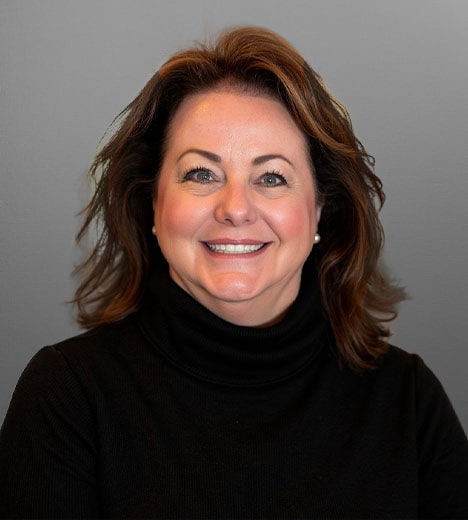 Headshot of a woman with shoulder-length brown hair, smiling warmly while wearing a black turtleneck, set against a gray background.