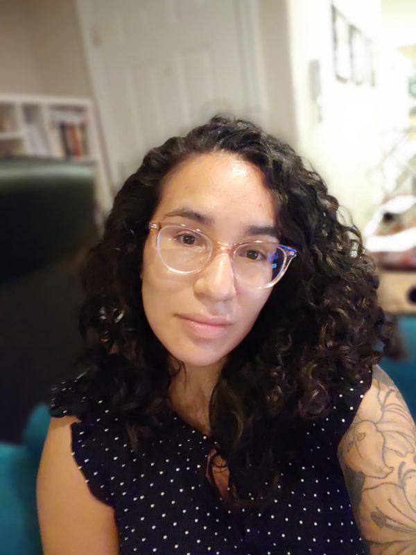 Maya has long wavy-curly brown hair and light brown skin and very cool glasses. She is wearing a polka dotted top with no sleeves and there is a hint of a beautiful linework tattoo on her shoulder