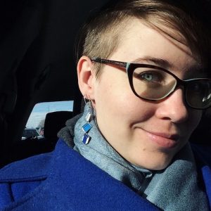 A profile pic of a person with very fair skin and stunning blue eyes wearing a blue jacket and scarf and terrific book earrings and glasses with short blonde hair
