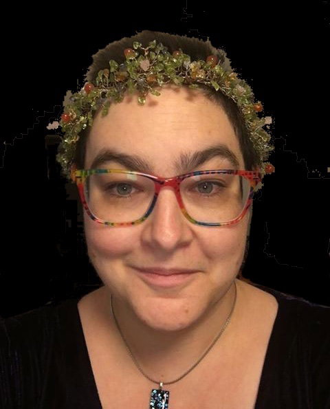 Catherine with red and multicolored glasses and a shaved head and a gorgeous green beaded headband. She is smiling and looking at the camera.