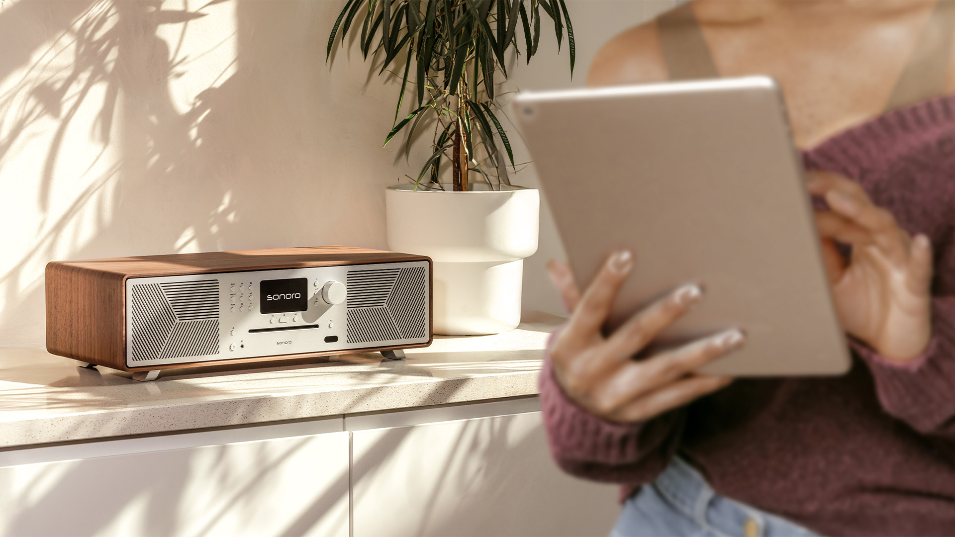 Sonoro MEISTERSTÜCK Musiksystem in Holzoptik auf einem Sideboard neben einer Pflanze, Person mit Tablet im Vordergrund.
