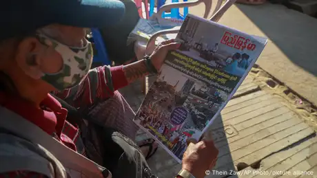 A man sits on a chair and looks at the front page of a newspaper.