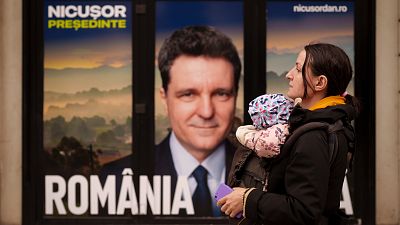 Uma mulher com um bebé ao colo passa junto a um cartaz de Nicusor Dan, candidato presidencial nas próximas eleições de maio, em Bucareste, 25 de março de 2025