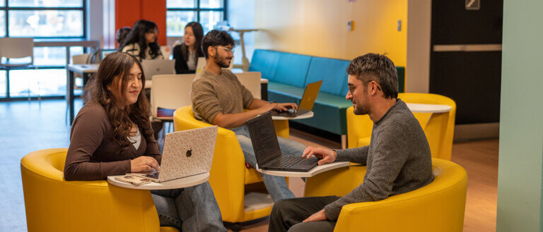 Four people work on laptops in a vibrant room with yellow chairs and natural light. They appear focused, creating a collaborative and productive atmosphere.