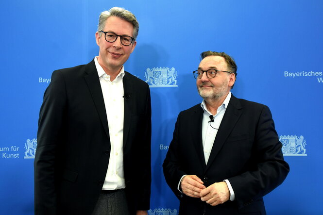 Markus Blume und Anton Biebl stehen vor einer blauen Wand mit dem bayerischen Landeswappen