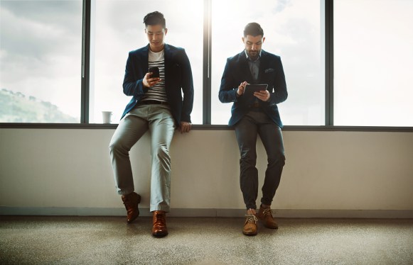 Shot of two young businessmen using a digital tablet and cellphone while leaning against a wall in a modern office