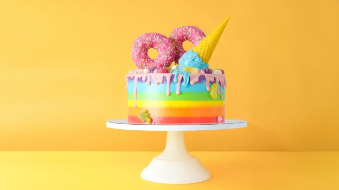 Cake on birthday with multicolored rainbow cream on a yellow background decorated with a waffle cone, pink donuts, sprinkles, marmalade candies poured with chocolate.