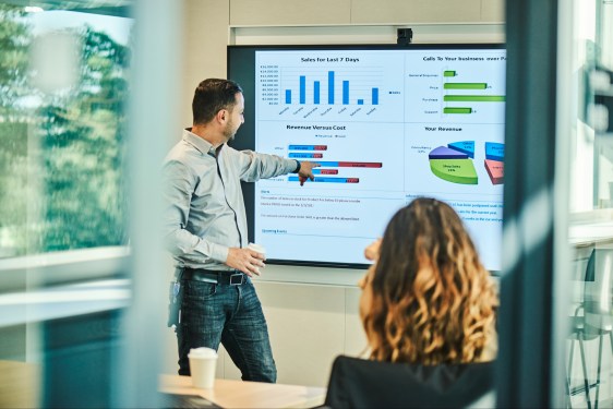 Presentation, man pointing to screen showing several types of graphs (bar, pie, etc.)
