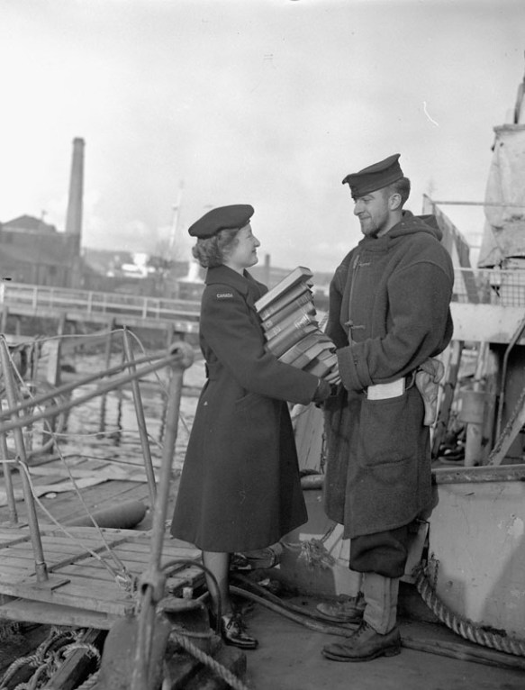 A black-and-white photograph of a member of the Women’s Royal Canadian Naval Service handing a man a tall stack of books beside a ship.