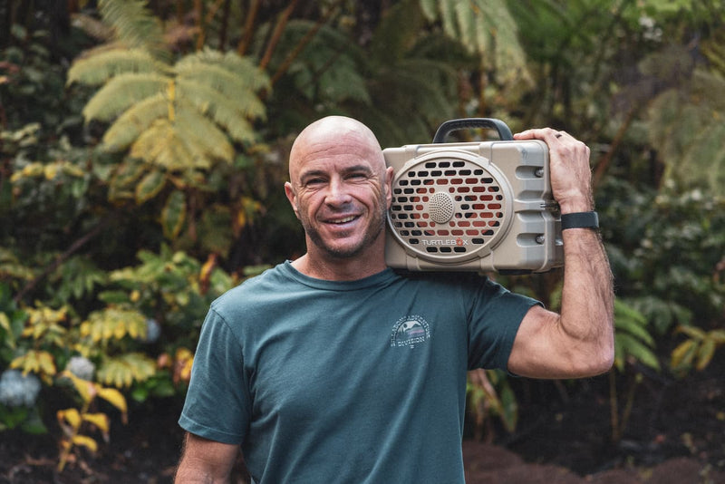 Turtlebox Ambassador, Shane Dorian, standing with a field Tan speaker on his shoulder in tropical woods.