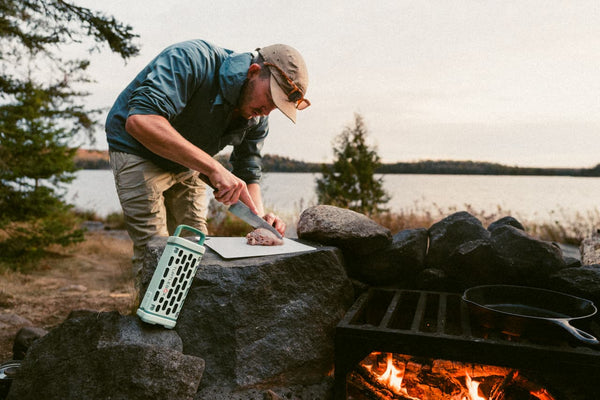 A lifestyle photo showing a man cutting meat next to an open camp fire with the River Rock Ranger speaker leaning against a rock. #base-color_river-rock