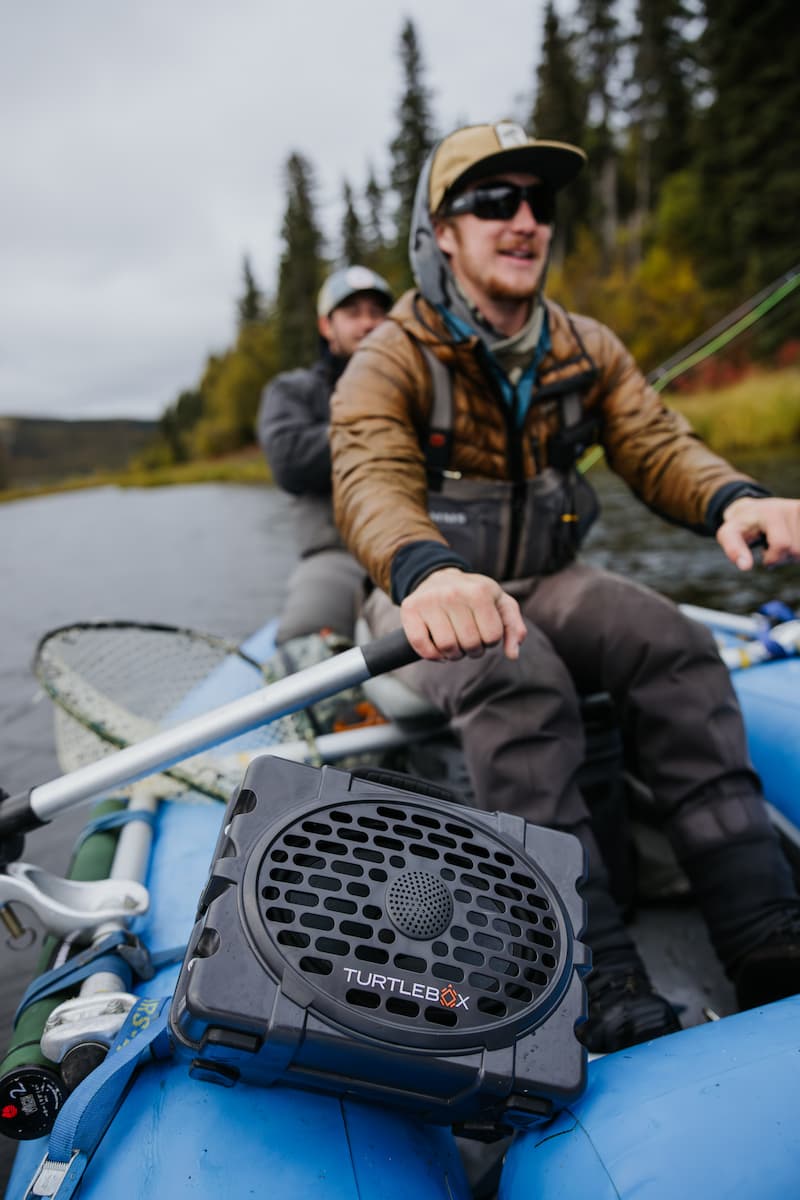 Guys rowing and fishing in a boat with the Turtlebox Original speaker in gray color attached to the boat
