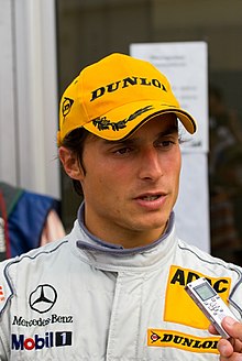 Photographie d'un homme brun, vu de face, en blanc avec une casquette jaune, répondant à des questions.