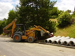 Un operaio attacca un cavo di sollevamento a una sezione di tubo fognario in cemento. Si notino gli stabilizzatori retratti di questo retroescavatore Case.