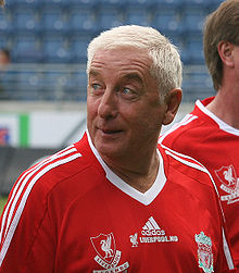 The head and upper torso of a gentleman in his late 50s. He has short white hair and is wearing a red football shirt, which has the Liverpool F.C. crest on the left breast, and a crest on the right breast that says "Liverpool Legends". A white logo of the Adidas sponsor is visible in the centre of the shirt, and three white stripes are present on the shoulder.