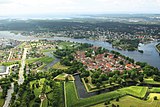 Luftbild einer an einem Fluss gelegenen Stadt. Zentral im Bild ist eine von einem sternförmigen Kanal vom Umland abgetrennten Insel.