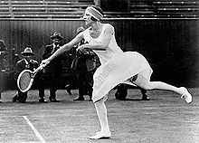 Photo en noir et blanc d'une femme en robe blanche à hauteur de genou, faisant un revers avec sa raquette de tennis.