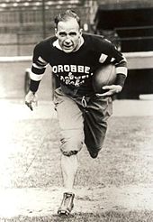 A football player in his mid thirties is seen running toward the camera with a ball tucked under his left arm. He balding, and wearing a dark coloured uniform with the words "Crosse and Blackwell Chefs" on his chest.