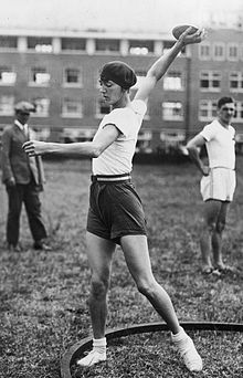 Photo en noir et blanc d'une femme vêtue d'un short sombre court et d'un T-Shirt blanc, le bras tendu, se préparant à un lancer de disque.