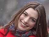 Una fotografía de Anne Hathaway; la cabeza se inclina hacia un lado y ella lleva un abrigo rojo con una bufanda púrpura.