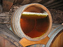 Photographie montrant une barrique à fond transparent révélant le voile de levure en surface sous lequel vieillit le vin jaune. Ces levures se présentent sous la forme d'une couche blanche irrégulière ; elles forment de petits stalactites qui s'enfoncent de quelques millimètres dans le vin dont on devine la couleur déjà sombre du vin jaune. Le mur en arrière plan est grisâtre, probablement revêtu de moisissures de chai.