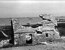 Au premier plan, une vieille maison de pierres en ruine ; au second plan, par les échancrures de ces ruines, la mer est recouverte de différents navires de guerres se dirigeant vers la plage.