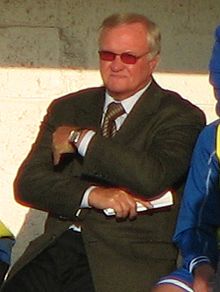 The upper body of a grey haired man sitting in the dugout with crossed arms. He has black trousers, a grey jacket, white shirt and a gold tie. He is wearing glasses with red tinted lenses a has a watch on his left wrist. In his right hand is a rolled up piece of paper.