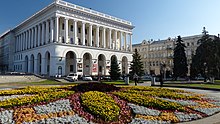 Farbfotografie mit Blick über ein rundes Blumenbeet zum klassizistisch erstellten Gebäude mit seinen weißen Säulen und Torbögen. Einige Tannen und ein weiteres Gebäude befinden sich auf der rechten Fotohälfte.