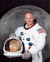 Un retrato de un hombre que llevaba un traje espacial, con su casco sobre la mesa frente a él. Detrás de él hay una gran fotografía de la superficie lunar.