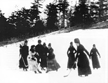 Photo en noir et blanc de femmes en robe et chapeau de fourrure jouant au hockey en extérieur.