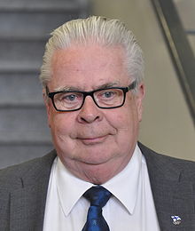 Late middle-aged man with gray hair combed back, wearing dark-rimmed eyeglasses, gray suitjacket, white dress shirt, and a blue necktie