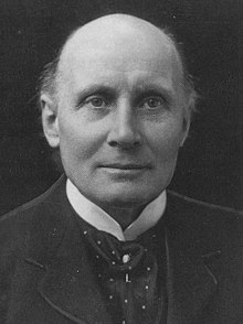 Photo showing Alfred North Whitehead from the front against a dark background looking at the camera, dressed in formal dark-colored attire with a high-collared white shirt below