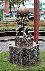 Zdjęcie pomnika w plenerze. Cztery nagie postacie unoszą glob złożony z niedokończonych puzzli. Symbol Wikipedii. Postacie stoją na postumencie wyglądającym jak równo ułożone stosy kartek lub książek.