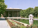 Water basin in a garden, flowers trees and a building with open portico.