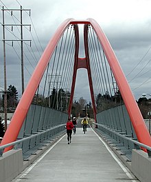 Two bicyclists and a jogger cross a long pedestrian bridge. The bright orange supporting arches of the bridge meet in the air about 30 feet (9 meters) above the bridge deck. Beyond the far end of the bridge is a paved bike path that continues into a woods in the distance.