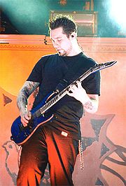 A man with brown hair is wearing a black T-shirt and black trousers while playing on a blue guitar. Tattoos are visible on both of his hands.