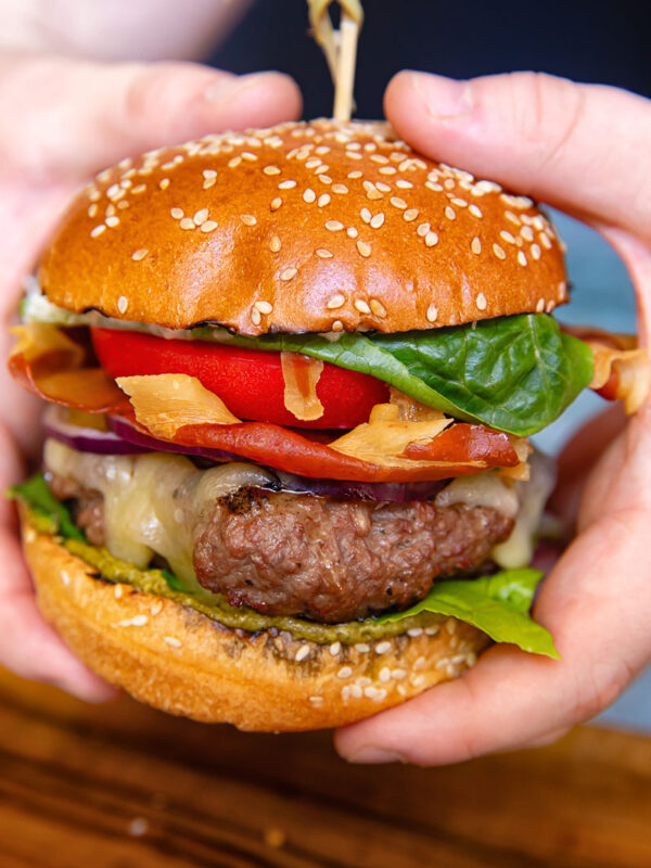 A vertical shot of a beef patty, tomato, lettuce and prosciutto in a bun in hands