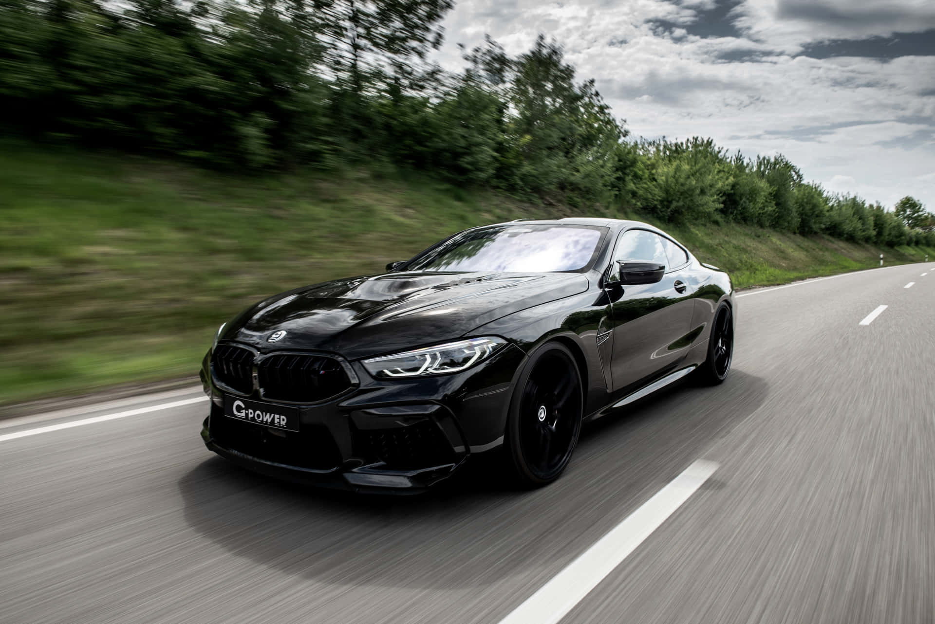 Schwarzes G-Power BMW 8er Coupé rast auf einer malerischen Straße mit grünen Bäumen und bewölktem Himmel. — Schwarze Sportwagen Wallpaper