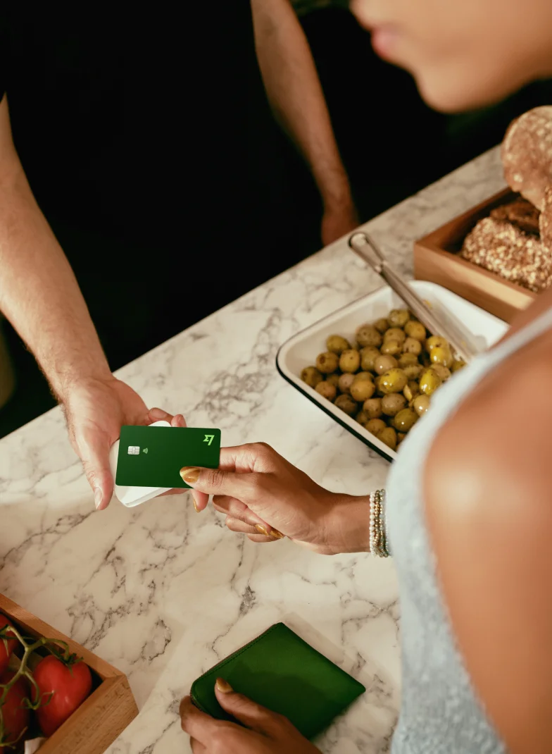Customer tapping a green card with the Wise logo onto a white contactless card reader over a counter.