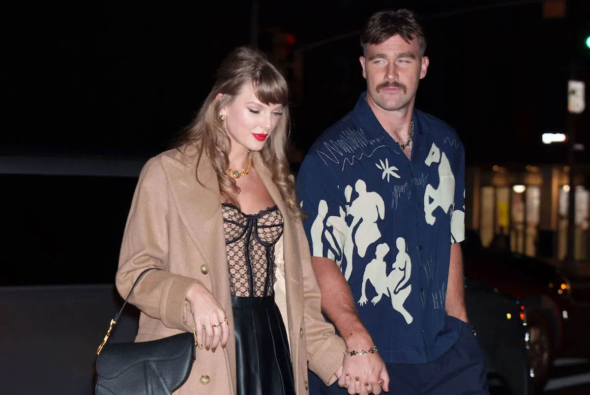 Taylor Swift looking down as she and Travis Kelce hold hands while walking at night. She's in a camel-colored jacket and he's in a blue and white short-sleeve button-up shirt.