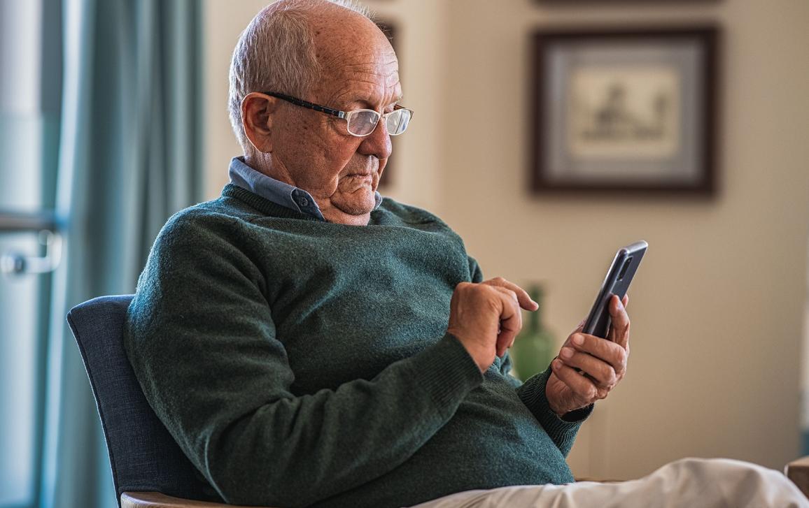 Starší muž sedící v křesle čtoucí obsah z mobilního telefonu