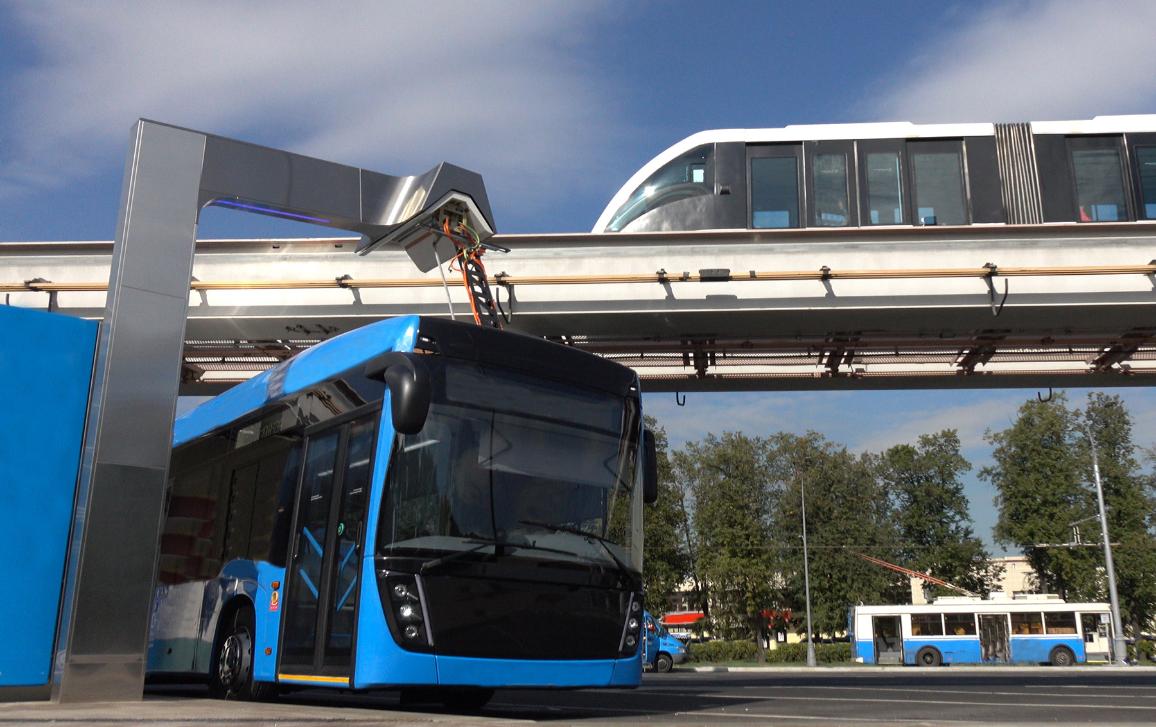 Un autobus elettrico blu alla stazione di ricarica mentre sullo sfondo passa un treno moderno sulla monorotaia