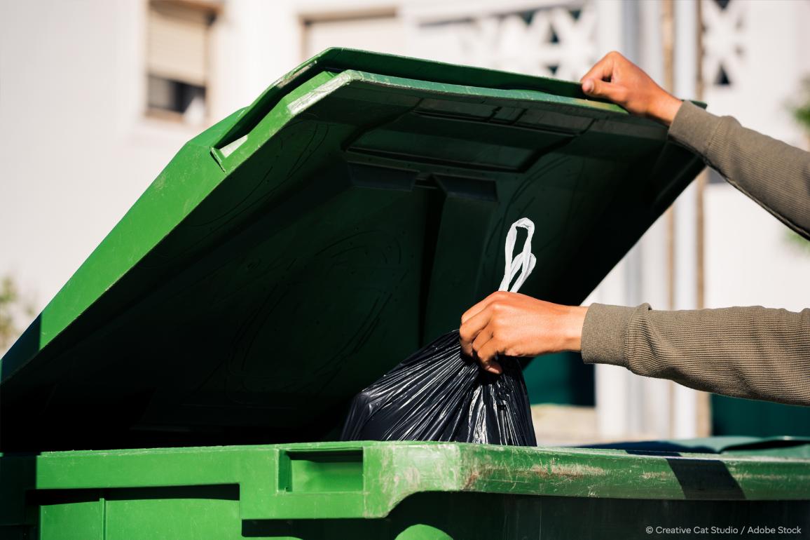 Slika, na kateri oseba meče črno vrečo za smeti v velik plastični zeleni zabojnik za smeti.