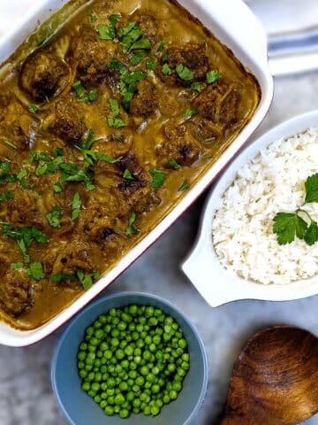 A dish of meatballs in curried onion sauce on a table next to a bowl of peas and a dish of rice.