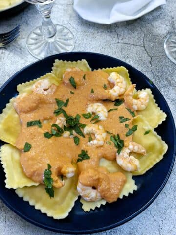 A blue dinner plate with pieces of ravioli topped with red pepper sauce and garnished with prawns.