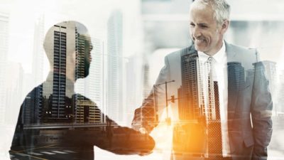 two businessmen shake hands amid a backdrop of tall buildings, indicating a share price movement or merger between ASX property companies