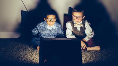 two young boys dressed in business attire and wearing spectacles sit side by side and watch closely an old fashioned television box receiver with built in wire ariels.