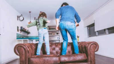 A dad and son dance on the sofa in their living room, wiggling their bottoms.