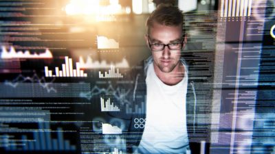 A man sits in casual clothes in front of a computer amid graphic images of data superimposed on the image, as though he is engaged in IT or hacking activities.