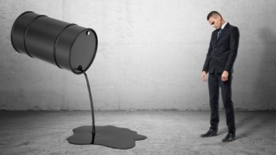 A man in a suit looks sad as oil is spilled from a barrel.